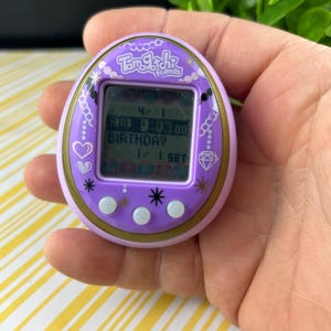 May include: A purple Tamagotchi Friends digital pet toy. The device has a small screen displaying text and numbers, with the word "BIRTHDAY" visible. The toy is held in a hand, with a yellow and white striped background.