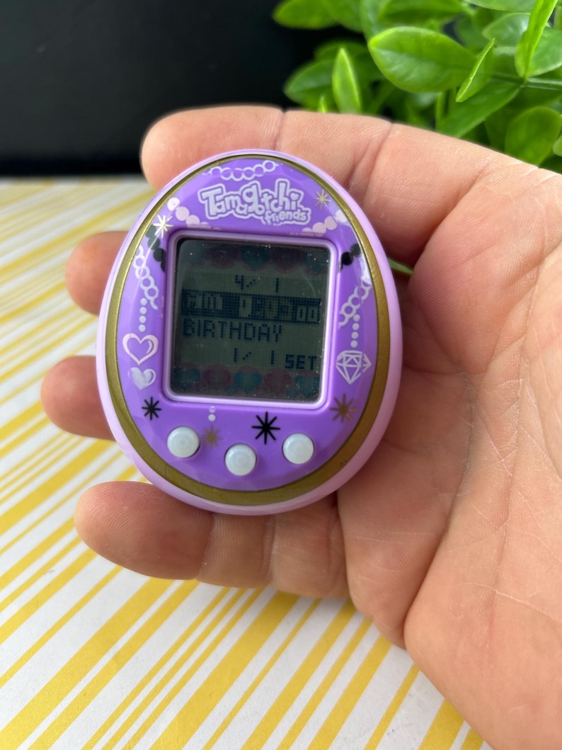 May include: A purple Tamagotchi Friends digital pet toy. The device has a small screen displaying text and numbers, with the word "BIRTHDAY" visible. The toy is held in a hand, with a yellow and white striped background.