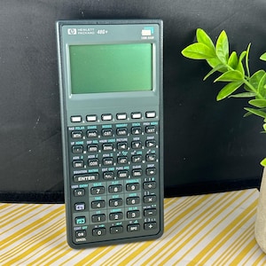 Könnte beinhalten: Dunkelgrauer Hewlett Packard 48G+ wissenschaftlicher Taschenrechner mit grünem Display und vielen Tasten. Der Rechner hat weiße und türkisfarbene Schrift. Er verfügt über 128K RAM und ist für fortgeschrittene mathematische Berechnungen konzipiert.