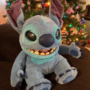 Puede incluir: Un peluche de Stitch azul con grandes orejas, una amplia sonrisa y ojos oscuros. El peluche está sentado sobre una superficie marrón, con un árbol de Navidad decorado al fondo.