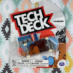 Puede incluir: Un fingerboard Tech Deck azul y negro con la palabra "Finesse" impresa en él. El fingerboard está sobre un fondo blanco y gris con un logotipo rojo y blanco. El embalaje incluye una etiqueta de advertencia para niños menores de 3 años.