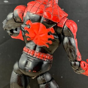 K&ouml;nnte beinhalten: Rote und schwarze Actionfigur eines Superhelden, von hinten gesehen. Die Figur hat ein rotes Spinnennetz-Design auf Kopf und Schultern sowie ein gro&szlig;es rotes Spinnenemblem auf dem R&uuml;cken. Der K&ouml;rper ist schwarz mit roten Akzenten.