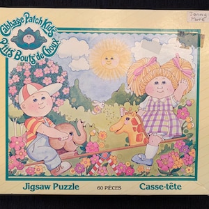 May include: A Cabbage Patch Kids jigsaw puzzle featuring two dolls on a seesaw. The girl doll is wearing a plaid dress and has blonde pigtails. The boy doll is wearing a striped shirt and a baseball cap. The puzzle has 60 pieces and is titled "Jigsaw Puzzle" and "Casse-tête".