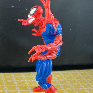 Peut inclure: Figurine articul&eacute;e de Spider-Man rouge, bleu et blanc. La figurine est dans une pose dynamique, avec un costume rouge et noir, des d&eacute;tails bleus et un masque aux yeux blancs. La figurine tire la langue.