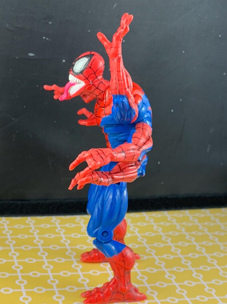 Peut inclure: Figurine articul&eacute;e de Spider-Man rouge, bleu et blanc. La figurine est dans une pose dynamique, avec un costume rouge et noir, des d&eacute;tails bleus et un masque aux yeux blancs. La figurine tire la langue.