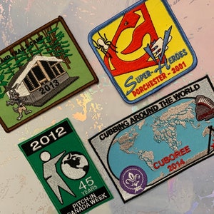 May include: Four embroidered patches with various designs and text. The patches are green, blue, yellow, and green. The text on the patches includes "Super-Heroes Dorchester-2001", "Cubbing Around the World", "Cuboree 2014", and "45 Years Pitch-in Canada Week".