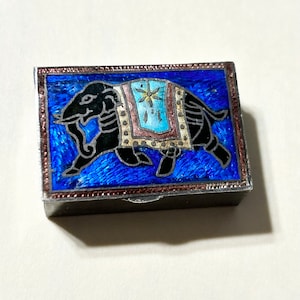May include: A small, rectangular silver box with a blue enamel top. The top features a black silhouette of an elephant with a colourful patterned saddle. The box has a silver border and a copper-coloured trim.