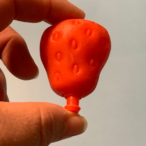 Peut inclure: Une fraise en plastique rouge avec une tige et un petit trou au fond. La fraise a une surface texturée pour ressembler aux graines.