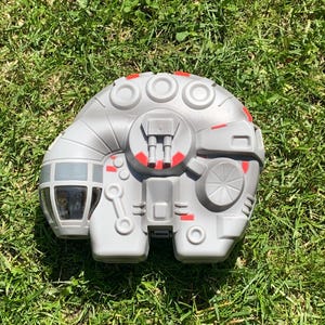 Op de afbeelding: Een grijs speelgoed ruimteschip met rode en grijze accenten, dat lijkt op de Millennium Falcon. Het speelgoed heeft een cockpit, cirkelvormige details en ligt op groen gras.