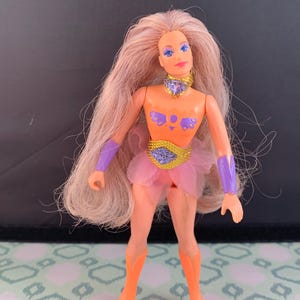 May include: A vintage action figure of a female character with long, light blonde hair. The doll wears an orange top with purple accents, a pink tutu, and orange boots. The figure has a purple choker and armbands, and a gold belt.