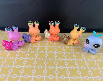 May include: A collection of colorful, small plastic animal figurines, including snails and a blue creature with large eyes. The snails are in shades of pink, orange, and yellow, with detailed shell designs. The figurines are on a yellow patterned surface.