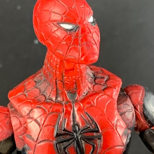 K&ouml;nnte beinhalten: Nahaufnahme einer rot-schwarzen Spider-Man-Actionfigur. Die Figur hat einen roten Kopf mit wei&szlig;en Augen und ein schwarzes Spinnennetz-Design. Die Brust zeigt ein schwarzes Spinnenemblem. Die Figur ist ein Sammlerspielzeug.