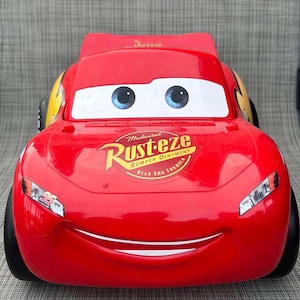 May include: A red toy race car featuring large white eyes with blue irises on the windshield. The hood displays a yellow and red Rust-eze Medicated Bumper Ointment logo. The front bumper shows the number 95 and a wide, smiling mouth.