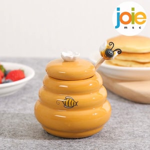 Può includere: Un vasetto di miele in ceramica gialla a forma di alveare, con un fiore bianco sul coperchio. Un piccolo misurino di miele in legno con un'ape sopra è all'interno. Il vasetto ha una grafica di un'ape dei cartoni animati sul lato. Il logo Joie msc è sullo sfondo.