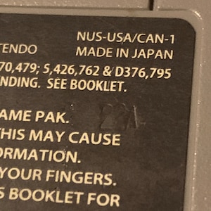 May include: Close-up of a Nintendo game cartridge label with the text "NUS-USA/CAN-1 MADE IN JAPAN 70,479; 5,426,762 & D376,795 INDING. SEE BOOKLET. AME PAK. THIS MAY CAUSE ORMATION. YOUR FINGERS. 5 BOOKLET FOR ERTISSEMENTS &Aacute;"