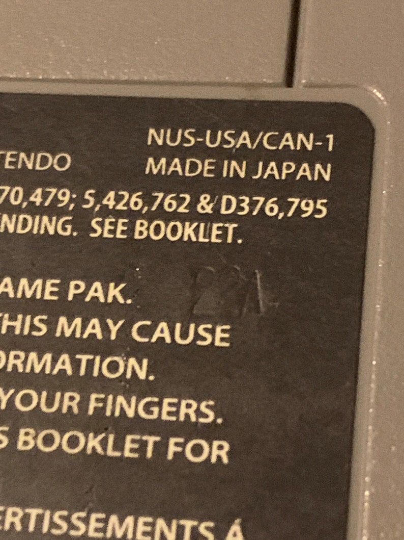 May include: Close-up of a Nintendo game cartridge label with the text "NUS-USA/CAN-1 MADE IN JAPAN 70,479; 5,426,762 & D376,795 INDING. SEE BOOKLET. AME PAK. THIS MAY CAUSE ORMATION. YOUR FINGERS. 5 BOOKLET FOR ERTISSEMENTS &Aacute;"