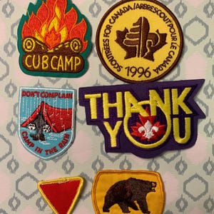 Op de afbeelding: Een verzameling geborduurde patches met diverse ontwerpen. Eén patch toont een kampvuur met de tekst "CUB CAMP". Een andere zegt "DON'T COMPLAIN CAMP IN THE RAIN". Andere patches bevatten een beer, een esdoornblad en de tekst "THANK YOU."