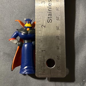 Pu&ograve; includere: Piccola action figure blu di un personaggio con volto giallo, corna e mantello rosso. La figura &egrave; alta circa 5 cm, misurata con un righello in acciaio inossidabile.