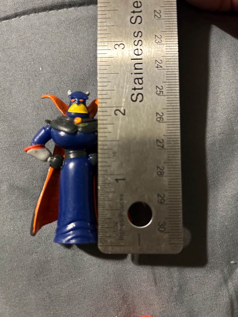 Pu&ograve; includere: Piccola action figure blu di un personaggio con volto giallo, corna e mantello rosso. La figura &egrave; alta circa 5 cm, misurata con un righello in acciaio inossidabile.
