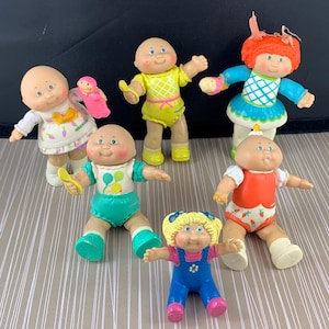 Op de afbeelding: Een verzameling van zes Cabbage Patch Kids poppen. De poppen zijn allemaal kaal met roze wangen en grote ogen. Ze dragen verschillende outfits, waaronder een gele jurk, een blauwe jurk, een groene romper, een rode romper en een blauwe overall.