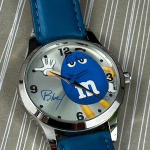 May include: A silver wristwatch with a blue leather strap. The watch face features a blue cartoon character with the letter "m" and the word "Blue" in script. The watch has silver hands and a silver bezel.