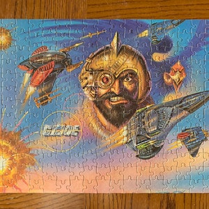 Peut inclure: Un puzzle coloré représentant une scène futuriste avec un homme portant un casque et un grand œil, un vaisseau spatial et une créature ressemblant à un oiseau. Le puzzle est intitulé "G.I. Joe".
