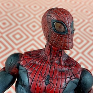 May include: A Spider-Man action figure with a red and black costume. The figure has a detailed web pattern on the red areas and a black spider emblem on the chest. The figure's mask has large eye openings.