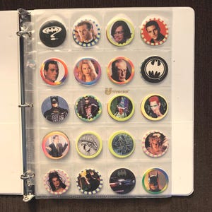 May include: A collection of Batman movie character pinback buttons in a binder. The buttons feature images of actors, the Batman logo, and other graphics. The buttons are in clear plastic sleeves.