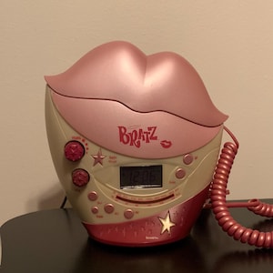 May include: A pink and beige Bratz-themed alarm clock shaped like a pair of lips. The clock features a digital display showing the time, along with buttons and a star design. The word "Bratz" is printed on the clock.