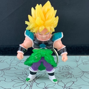 May include: A yellow-haired anime character figurine with a green and purple outfit. The figurine is holding a key chain.