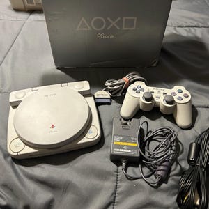 May include: A light gray Sony PS one game console with a matching controller, a power adapter labeled 7.5V, and a black AV cable. The console is positioned in front of its original gray box, which features the PlayStation symbols and the PS one logo.