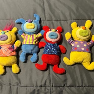 Op de afbeelding: Vier kleurrijke pluche speelgoed met grote, ronde gezichten en open armen. Elk heeft een ander kleurenschema: geel met roze haar, blauw met gele strepen, rood met blauwe stippen en geel met paarse stippen. Ze hebben grote, ronde ogen en open monden.