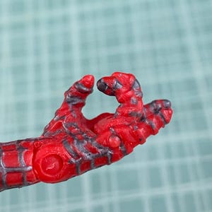 May include: Close-up of a red and black action figure hand. The hand is in a gesture, with the fingers slightly curled. The figure's hand has a textured appearance with detailed lines and patterns.