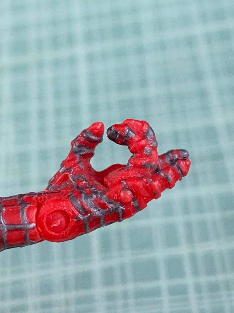 May include: Close-up of a red and black action figure hand. The hand is in a gesture, with the fingers slightly curled. The figure's hand has a textured appearance with detailed lines and patterns.