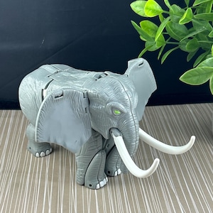 以下が含まれることがあります： 大きな白い牙と緑の目を持つ灰色の象のおもちゃ。おもちゃには関節があり、プラスチック製です。象の耳は明るい灰色の色合いです。おもちゃは、白い線のある明るい茶色の表面にあります。