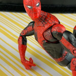 K&ouml;nnte beinhalten: Eine rot-schwarze Spider-Man-Actionfigur. Die Figur hat ein detailliertes Design mit roter Maske und Anzug, schwarzen Akzenten und beweglichen Gelenken. Die Figur ist mit einem ausgestreckten Arm posiert.