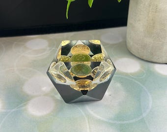 May include: A geometric ashtray made of clear resin with gold coins embedded inside. The ashtray has a dark grey exterior. It is on a light green and white surface. A small potted plant is in the background.