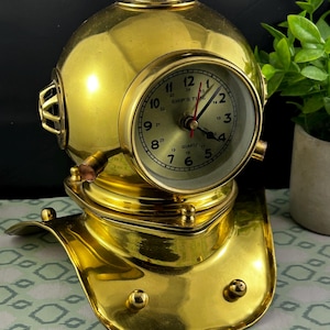 May include: A brass-colored desk clock shaped like a vintage diving helmet. The clock face is round with black numerals and hands, and the words "SHIP'S TIME" and "QUARTZ" are visible. The helmet has intricate details and a polished finish.