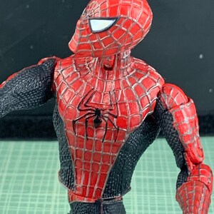May include: A Spider-Man action figure, featuring a red and black costume with a webbed design. The figure has a detailed mask with white eye lenses and a black spider emblem on the chest. The figure is posed with its arm extended.
