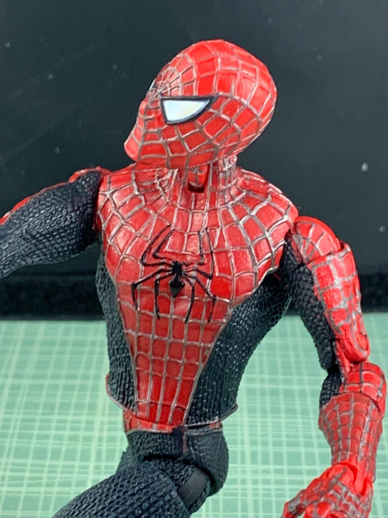 May include: A Spider-Man action figure, featuring a red and black costume with a webbed design. The figure has a detailed mask with white eye lenses and a black spider emblem on the chest. The figure is posed with its arm extended.