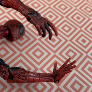 May include: A red and black Spider-Man action figure, featuring detailed textures on the arms and hands. The figure's design includes a textured pattern, with a focus on the articulated limbs. The figure is set against a patterned background.