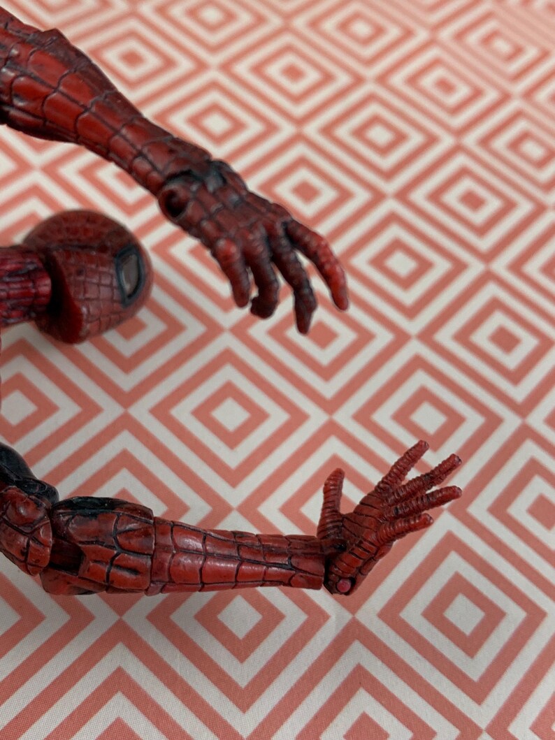 May include: A red and black Spider-Man action figure, featuring detailed textures on the arms and hands. The figure's design includes a textured pattern, with a focus on the articulated limbs. The figure is set against a patterned background.