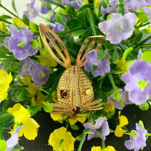 Op de afbeelding: Een gouden konijnen broche met rode strass steentjes als ogen. De broche is omringd door paarse en gele bloemen.