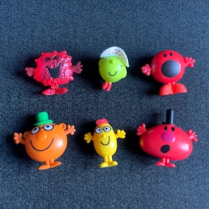 May include: Six colorful Mr. Men and Little Miss figurines. The characters are in various colors, including red, orange, yellow, and green, with distinct facial features and accessories like hats and glasses. They are arranged on a dark blue surface.