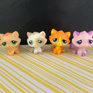May include: Four small, colorful cat figurines. The cats are in shades of tan, white, orange, and pink, each with large, expressive eyes and small paws. They are arranged on a yellow and white striped surface.