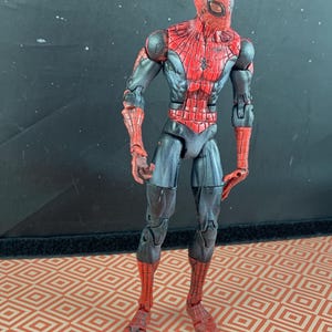 May include: A Spider-Man action figure in a standing pose. The figure is predominantly red and black, with a detailed web pattern on the red sections. The figure has a spider emblem on the chest and is on a patterned surface.