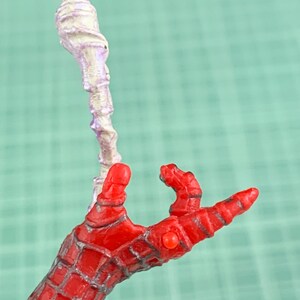 May include: A red and gray Spider-Man action figure hand with a white and purple object. The hand is in a web-like pattern. The object is a twisted, bulbous shape.