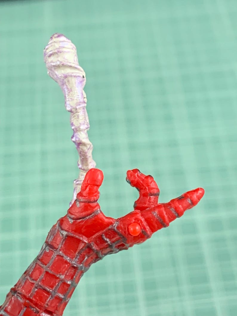 May include: A red and gray Spider-Man action figure hand with a white and purple object. The hand is in a web-like pattern. The object is a twisted, bulbous shape.