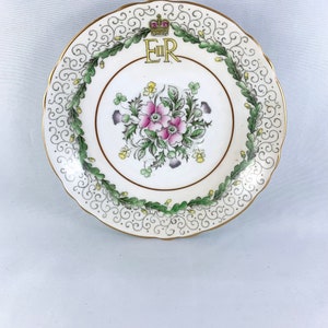 K&ouml;nnte beinhalten: Ein wei&szlig;er Porzellanteller mit goldfarbenem Rand und einem gr&uuml;nen und goldenen Blumendesign. In der Mitte des Tellers befindet sich eine goldene Krone &uuml;ber den Buchstaben "ER".