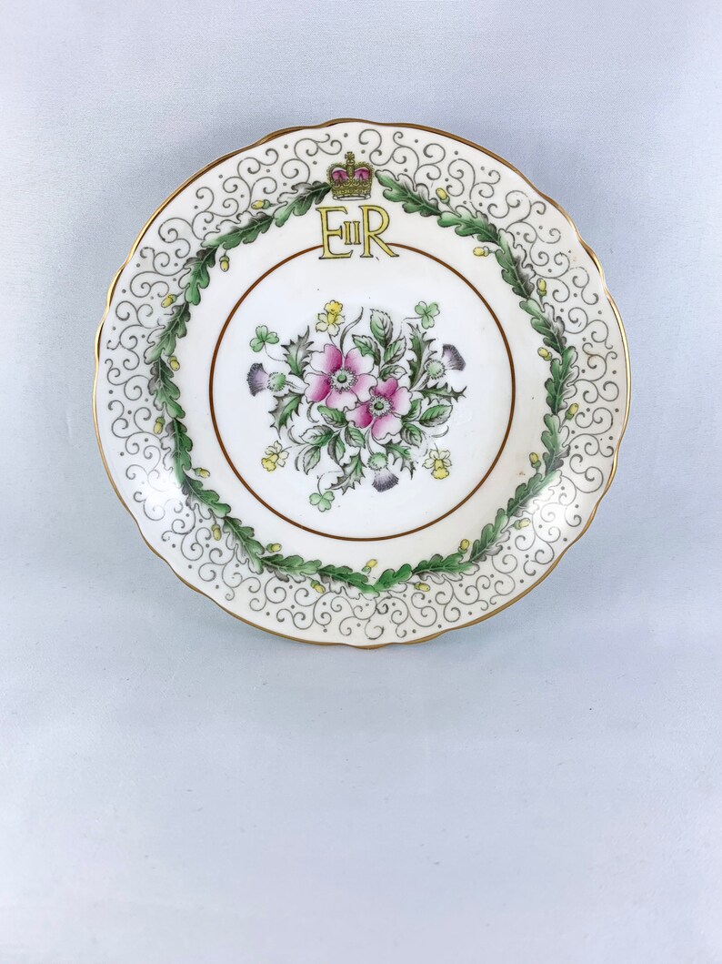 K&ouml;nnte beinhalten: Ein wei&szlig;er Porzellanteller mit goldfarbenem Rand und einem gr&uuml;nen und goldenen Blumendesign. In der Mitte des Tellers befindet sich eine goldene Krone &uuml;ber den Buchstaben "ER".